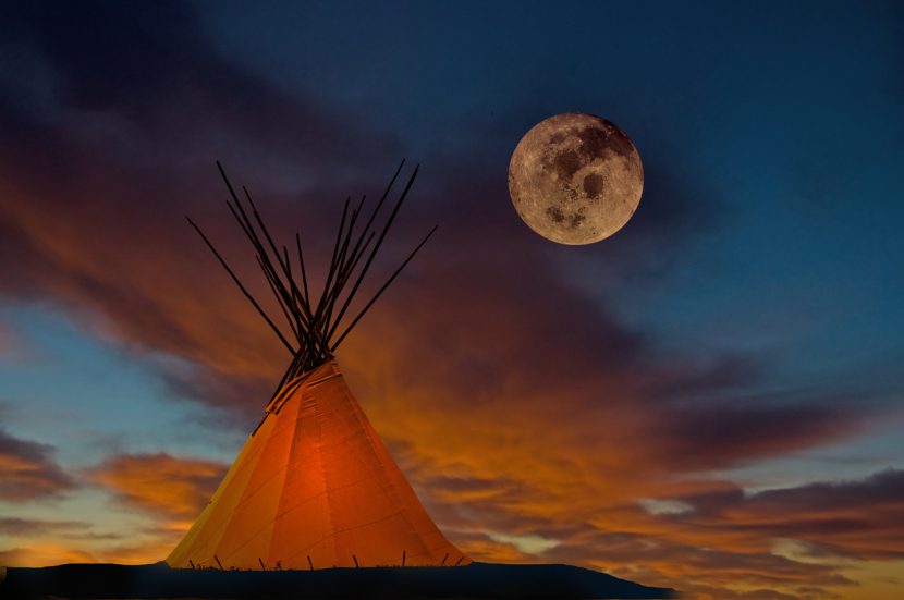A prairie First Nation teepee with interior light at sunset. Full moon